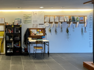 retrofitted lab and library space in CAPA with shelving, a carroll, and cork boards against a white wall in a hallway; an assortment of books, scientific instruments, lab and field materials, including organic matter, are collected on the wall and furniture