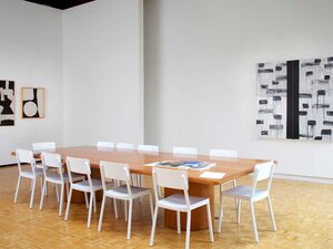 white room with rectangular wooden table surrounded by white chairs with black and white paintings on the wall
