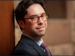 slightly smiling man in a suit with tortoiseshell glasses leans against a doorway