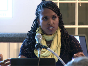 Woman with braids and a yellow scarf lecturing at a microphone