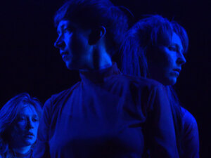 three women stand, looking in different directions, bathed in blue light