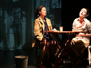Two women on stage sitting across a table from each other (still from the production)