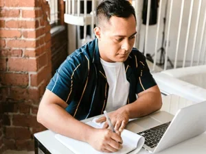 a student sits at a table while researching on a laptop and taking notes on a notepad