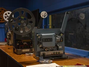 A side view of two reel-to-reel film projectors on a counter in the projection room of a theater. They are pointed toward the screen. 