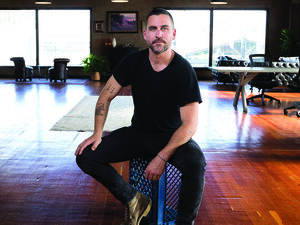 Bryn Mooser: A man sits on a chair in a room with a wooden floor wearing a black tee shirt and looking directly at camera