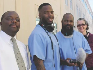 three men pose for the camera, two wearing blue, one with headphones, and one in white