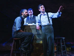 Three actors in period dress (suspenders, etc.) on stage pointing 