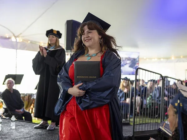 Female student with long brown hair wearing cap and gown with a red dress and turquoise necklace hugs her diploma as she walks in front of stage back to her seat