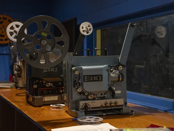 A side view of two reel-to-reel film projectors on a counter in the projection room of a theater. They are pointed toward the screen. 