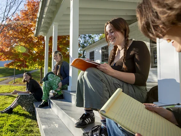 Four students smiling and sitting with notebooks on the porch of Cricket Hill in autumn. 