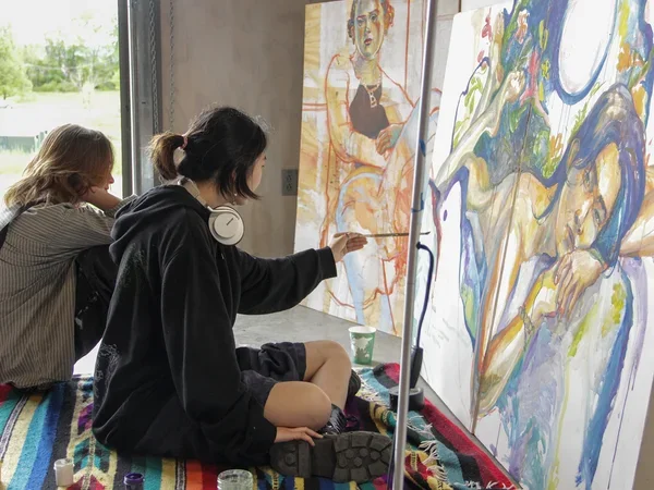 student wearing a black hooded sweatshirt sitting cross-legged on the floor and painting a figure on a large horizontal canvas
