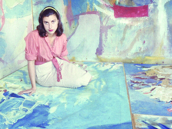 Young woman with brown hair, pink blouse and white skirt sits on a painting with paintings in background 