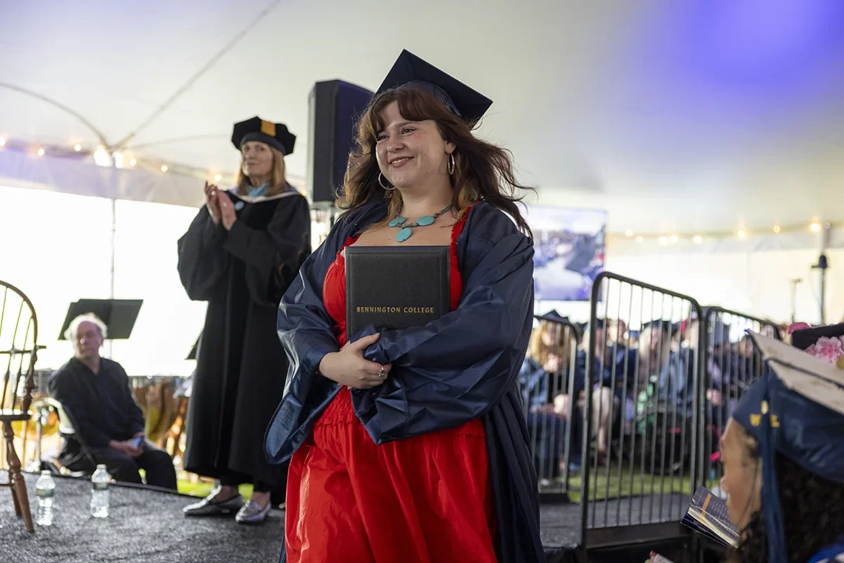Female student with long brown hair wearing cap and gown with a red dress and turquoise necklace hugs her diploma as she walks in front of stage back to her seat