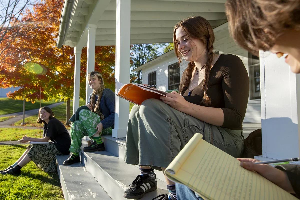Four students smiling and sitting with notebooks on the porch of Cricket Hill in autumn. 