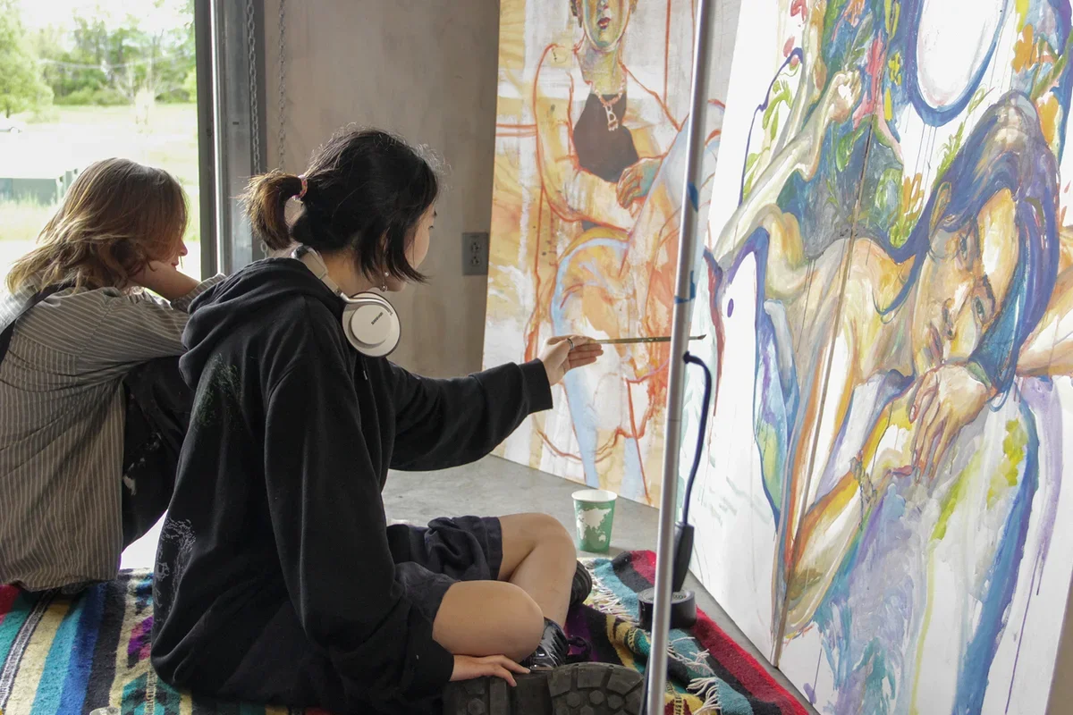 student wearing a black hooded sweatshirt sitting cross-legged on the floor and painting a figure on a large horizontal canvas