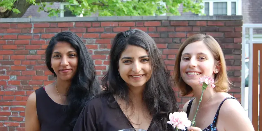 three students in a garden in fancy dress hold cocktails and smile at the camera