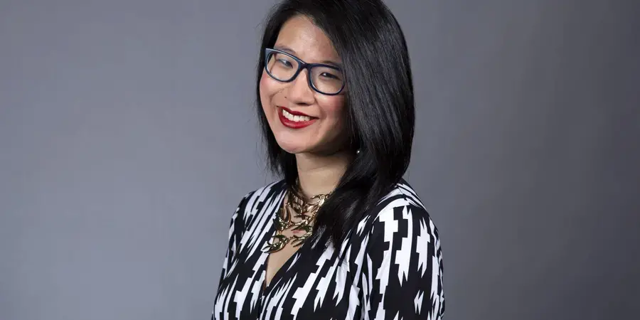 Dark haired woman wearing glasses and black and white blouse against grey background