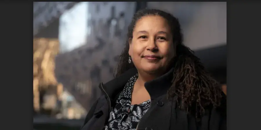 woman with a half-smile and a black coat is in focus against a blurred-out cityscape background