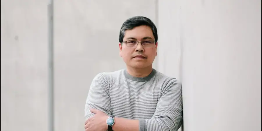 man in a striped shirt leans against a white exterior wall