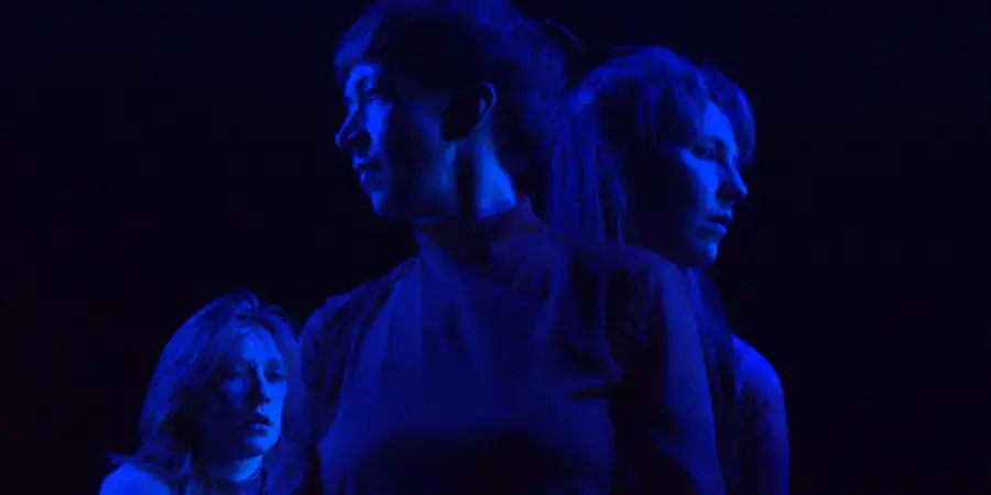 three women stand, looking in different directions, bathed in blue light