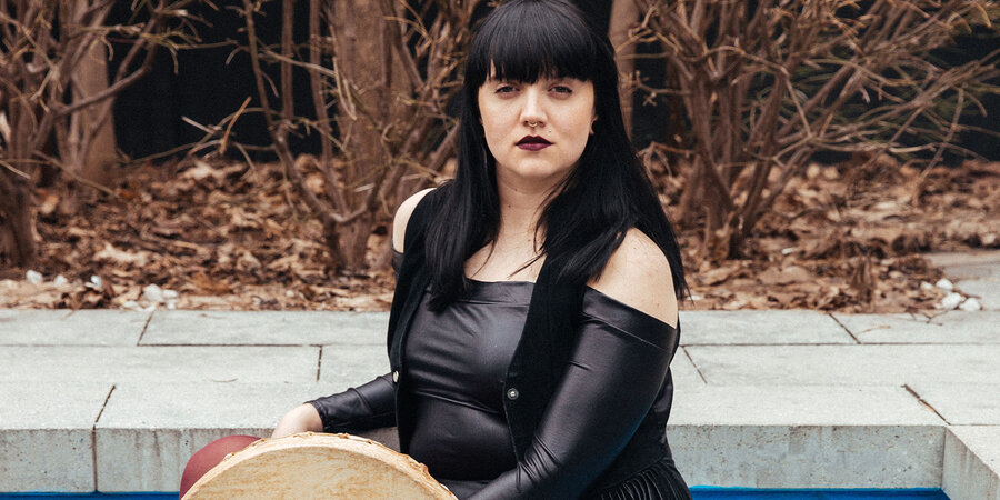woman with black hair and a shiny dress holds a skin drum in what looks like a swimming pool