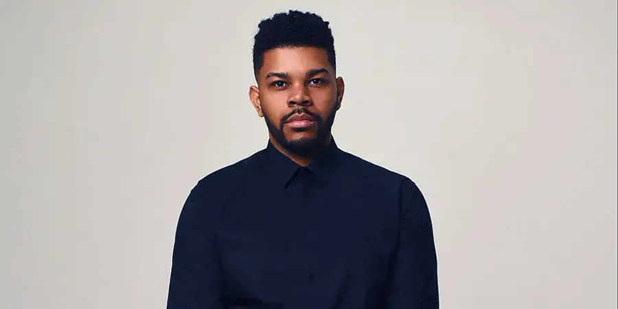 A serious-looking man with brown skin, dark hair, who is wearing a dark blue button up shirt.