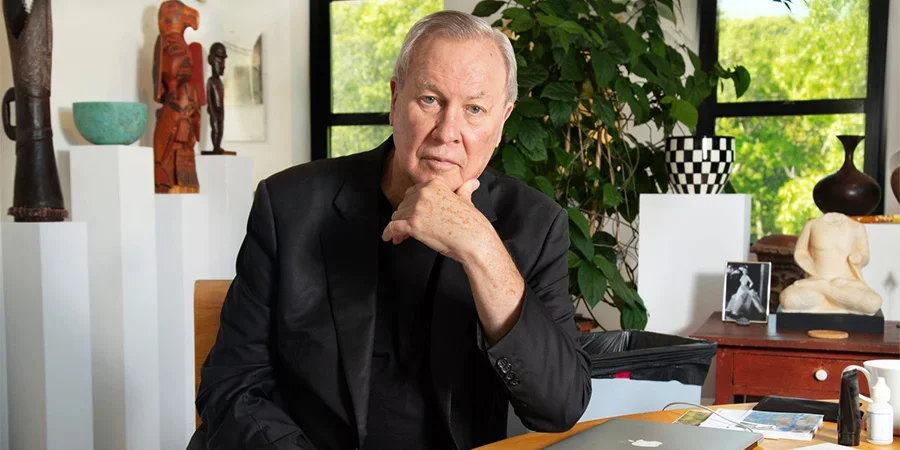 Robert Wilson, director and artist, seated at a desk with sculptures and plants in a well-lit room.