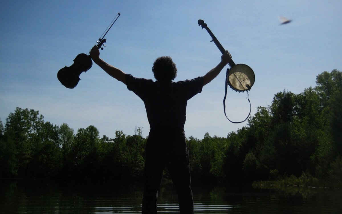 A man (seen from behind) stands with his feet in a lake on a sunny day holding two music instruments