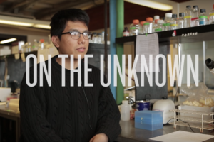 student gazing off into the distance seated in a laboratory, with text that says "On the Unknown" across the screen