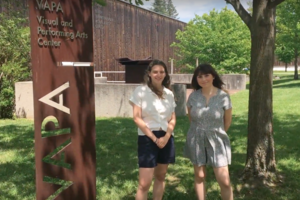 two students in summer clothes beside metal "VAPA" sign with grass, trees, and the flank of a wood-sided building