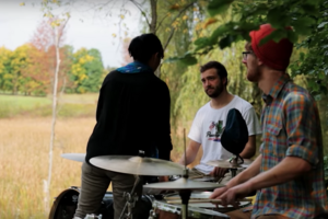 three people and two drum kits, outdoor drum class in the shade with a sunny field of high grass on the left