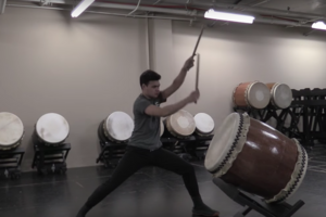 man raises arms to strike a Taiko drum in a florescent-lit windowless room with drums stacked against the cement wall