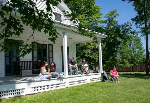 students sitting on a shady porch of a white clapboard house on a sunny day