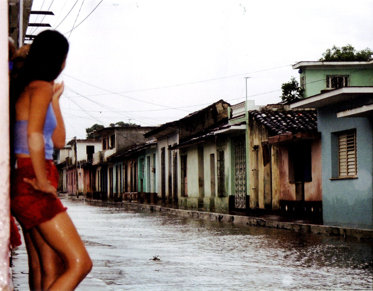 Woman wearing red shorts looks down an empty street of faded, colorful houses during a rainstorm in Cuba
