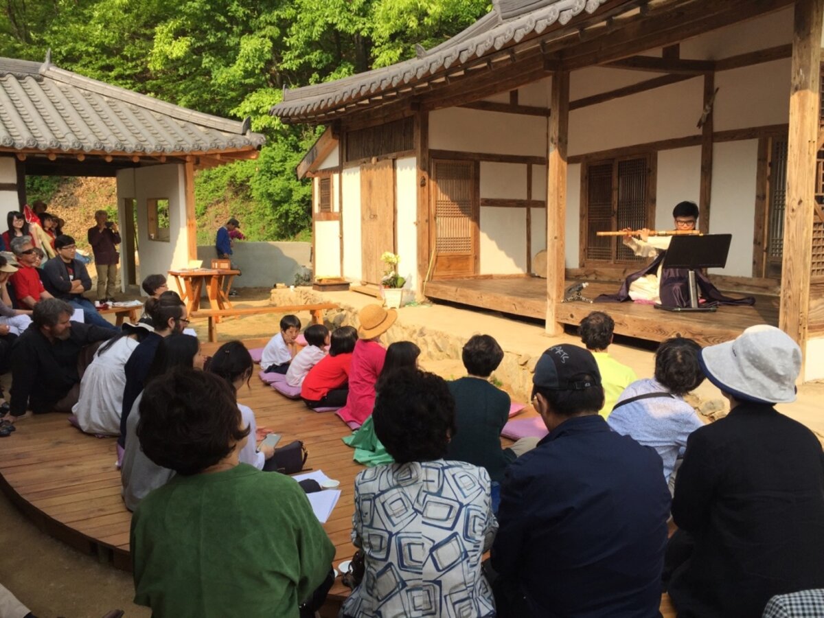 A musician plays the flute on the open air porch of the traditional Korean house in front of a seated audience