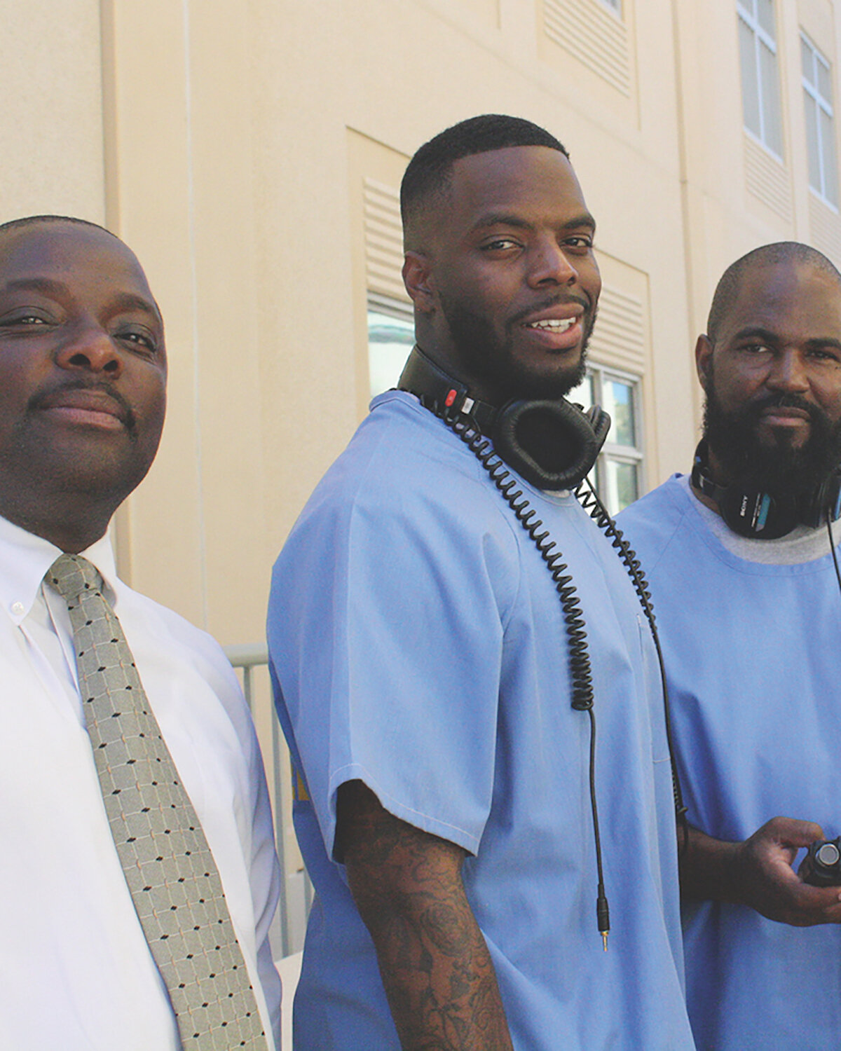 three men pose for the camera, two wearing blue, one with headphones, and one in white