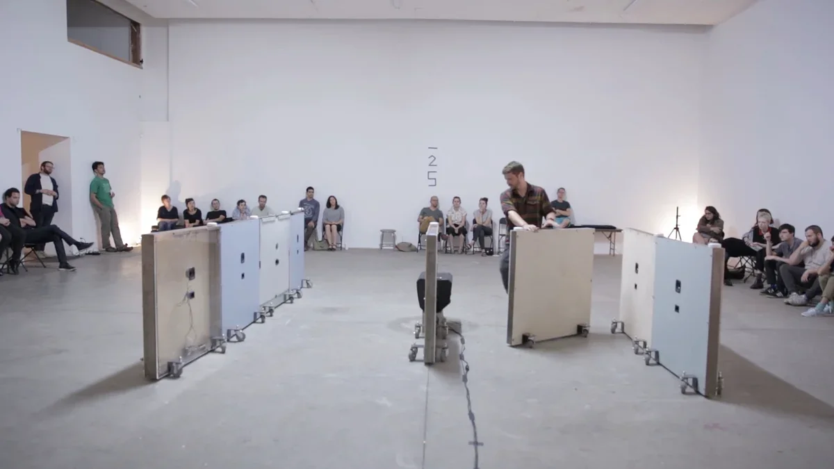An artist moves one of the white, wheeled, half-size walls around the performance space while the audience watches from the sides
