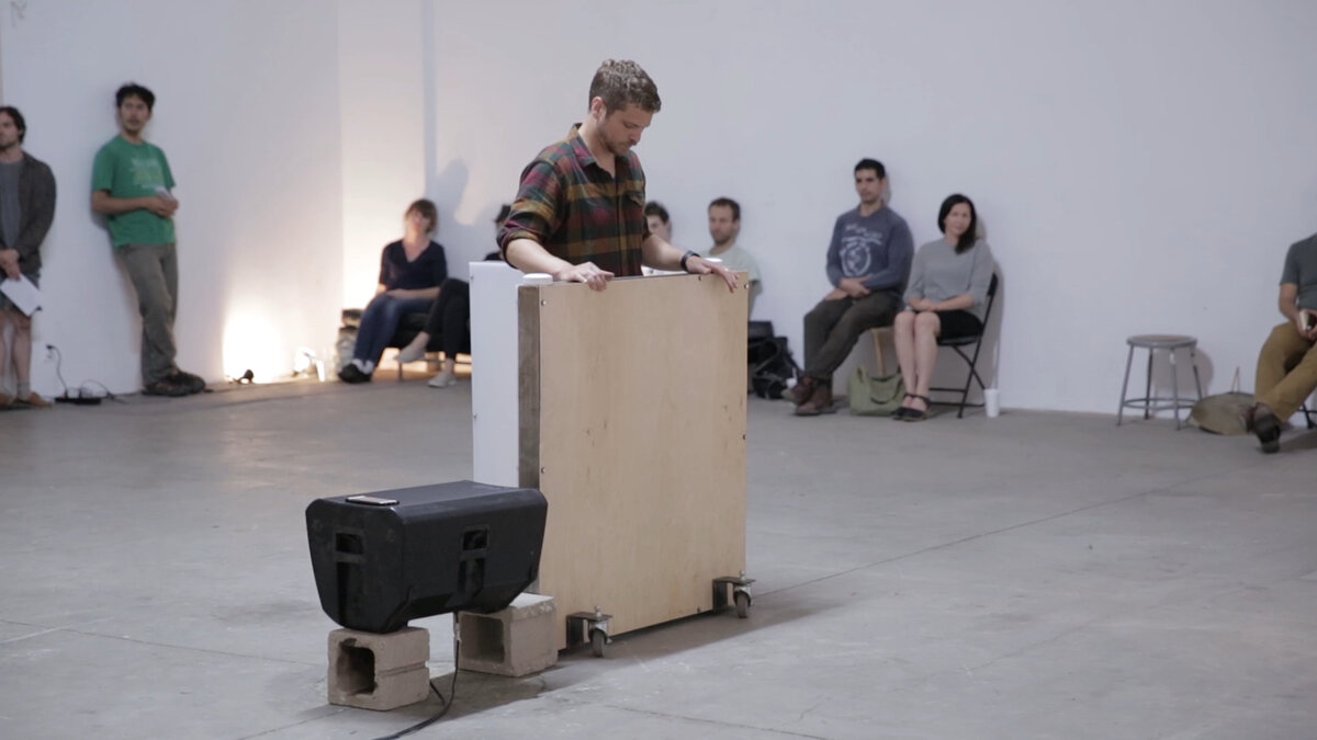 An artist moves one of the white, wheeled, half-size walls around the performance space while the audience watches from the sides