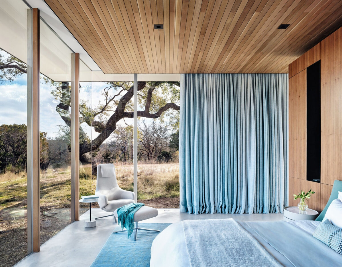 A bedroom with blue curtain, rug, and bedspread and glass walls that show a tree outside