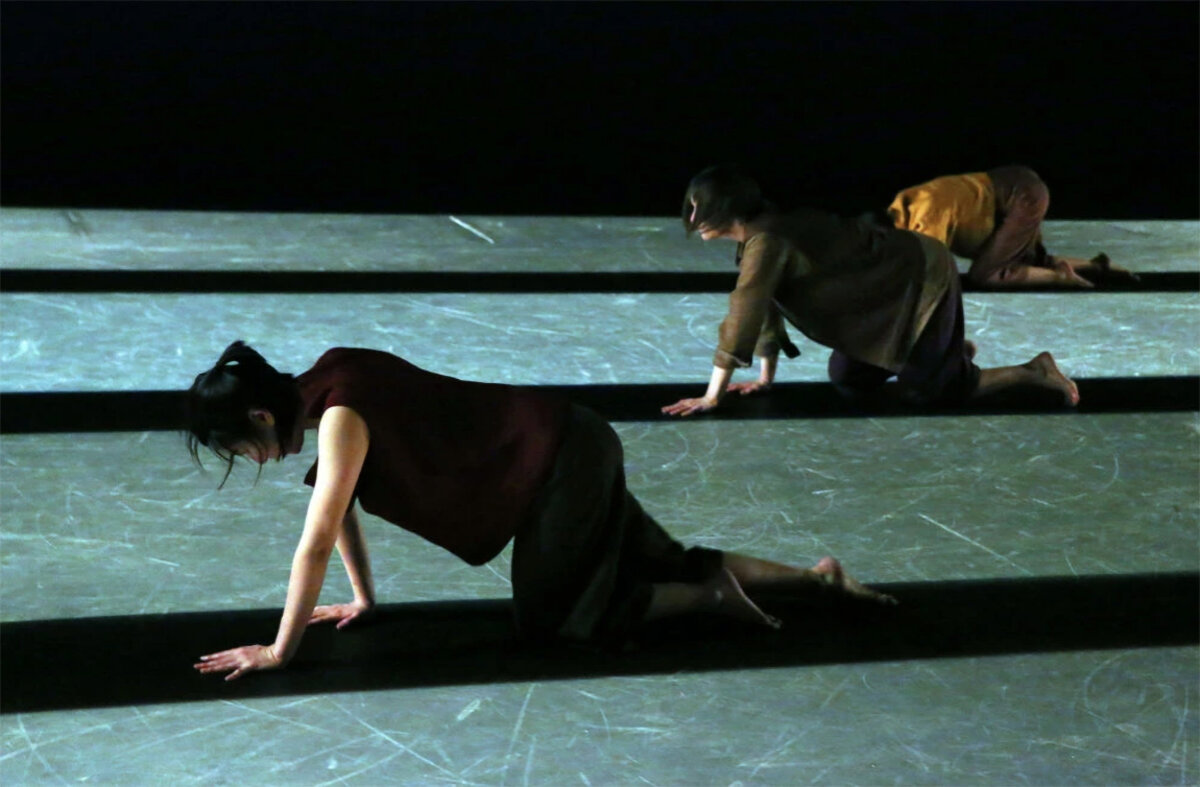 Three dancers crawl on all fours on three black lines on the ground