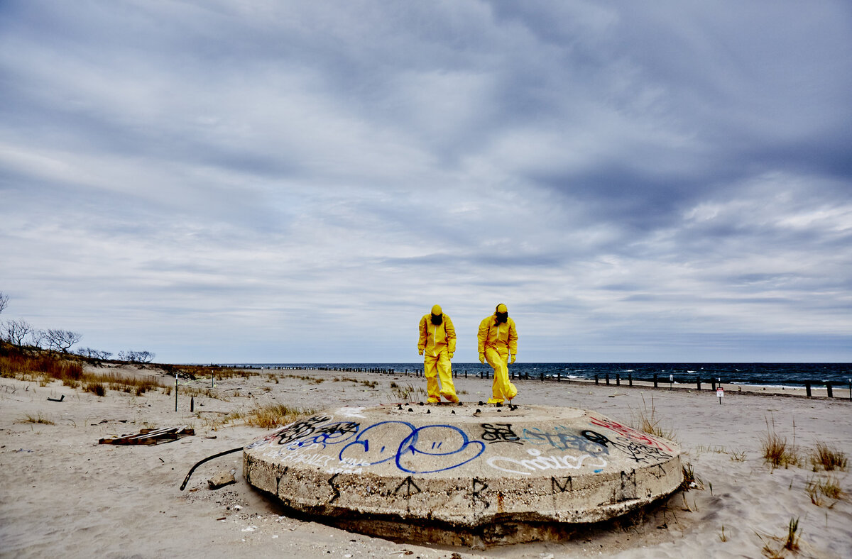 Two people in yellow hazmat suits stand on top of a graffiti-covered concrete structure on the beach