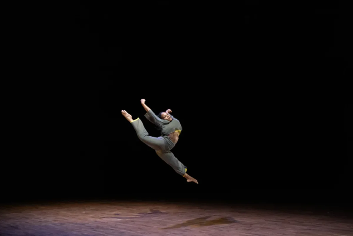 A dancer in gray performs a high split leap against a black stage background.