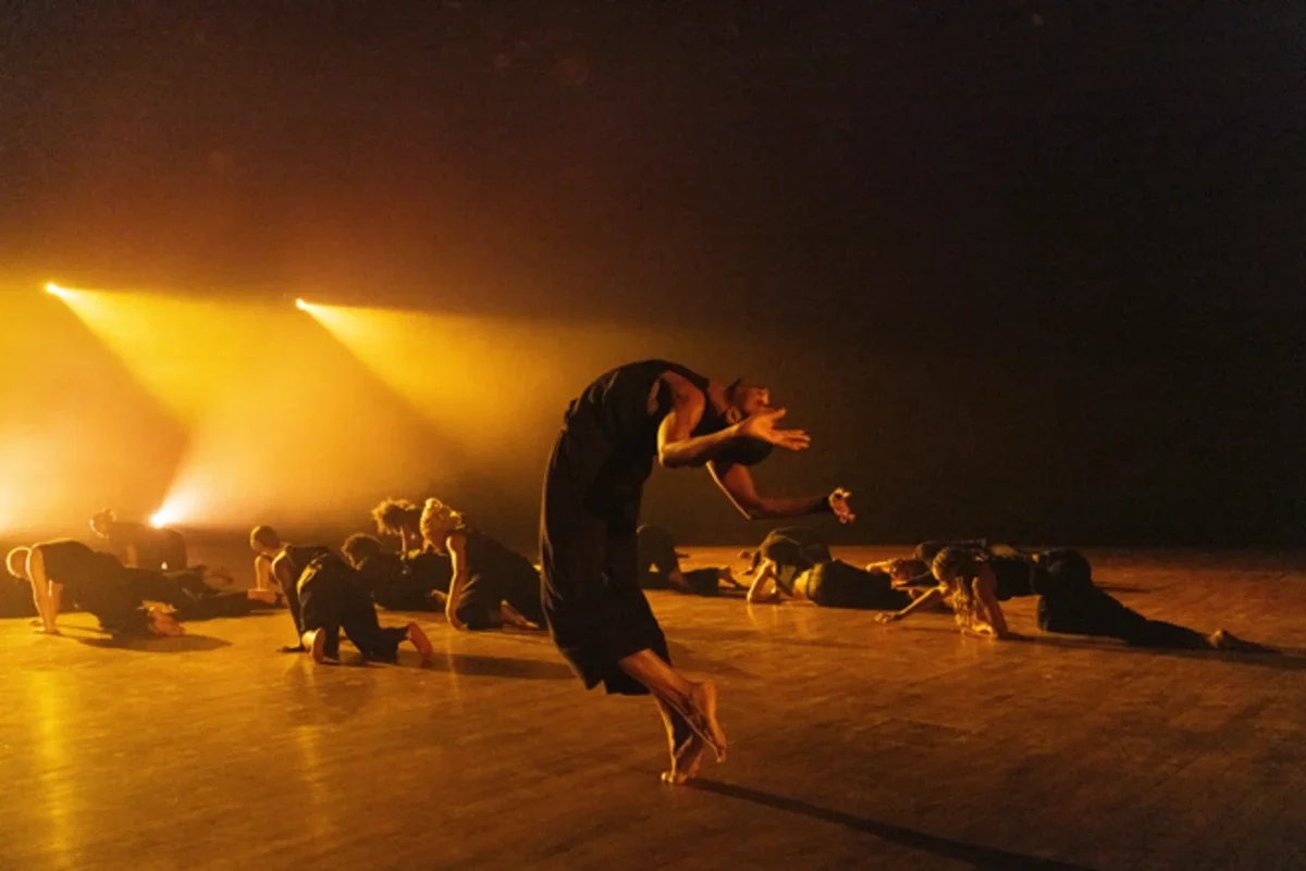 A dancer arches backward with hands raised near their face as other performers move across the floor under golden stage lights.