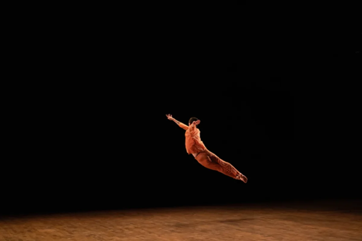 A dancer dressed in orange extends horizontally through the air above a dark stage.