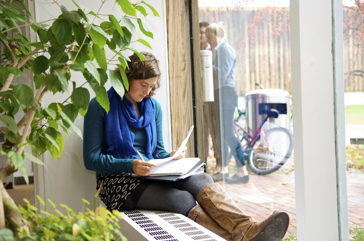 student writing in a notebook while sitting on a windowsill behind a green plant