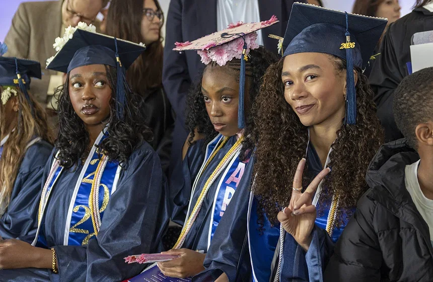 A row of seated graduates with decorated caps. The right most grad looks directly at the camera and gives a peace sign with her fingers.