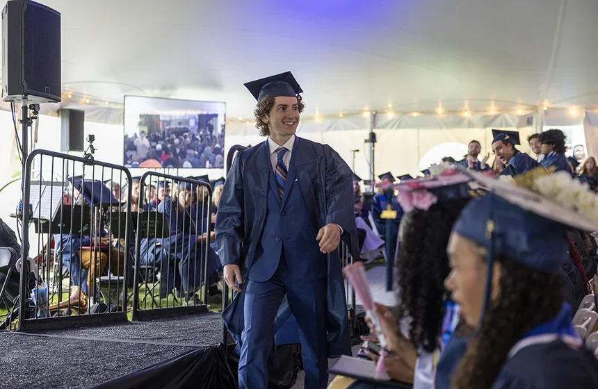 Handsom male student in blue suit, cap, and gown looks into the audience as he crosses in front of the stage