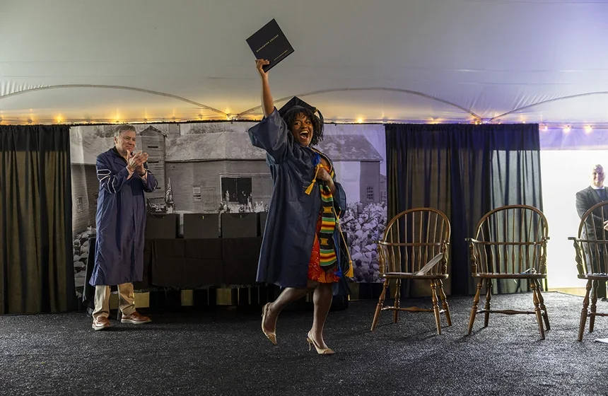 Excited female student in a brightly colored dress, cap, and gown holds her diploma high overhead while Nick Stephens '77, chair of the Board of Trustees, claps