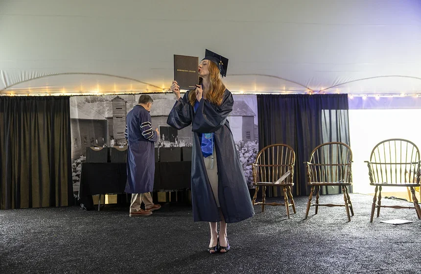 On stage, a. female student holds her diploma to the side and kisses its edge dramatically