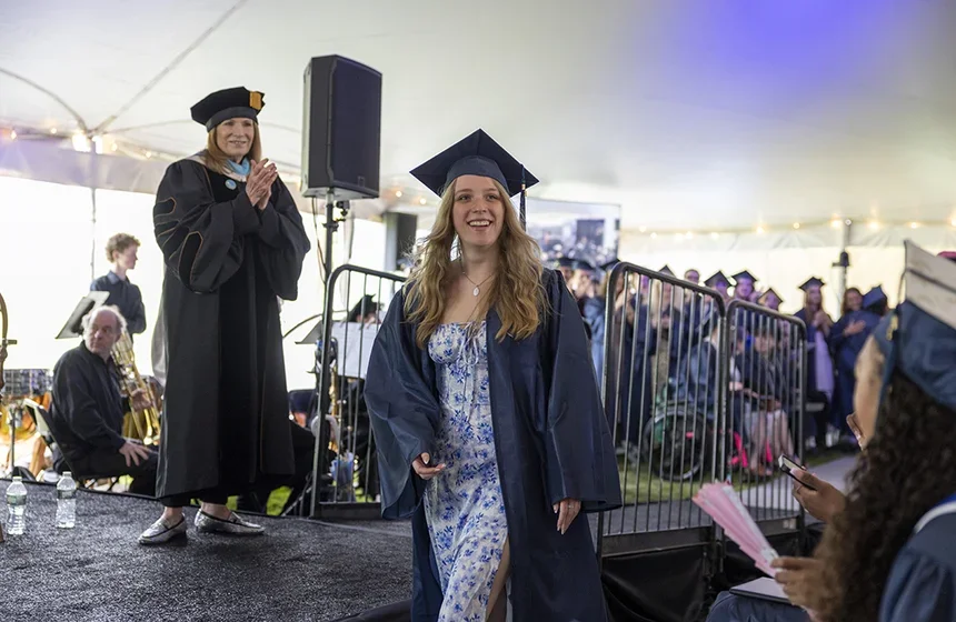 female student with long wavy blond hair in blue flowered dress, cap, and gown walking in front of stage on graduation day